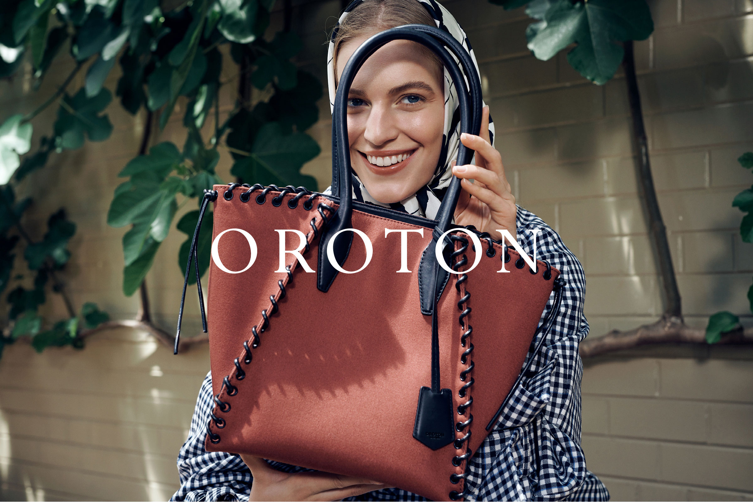 Smiling woman holding a terracotta purse with black trim. She is wearing a black and white checkered scarf and has green foliage behind her.