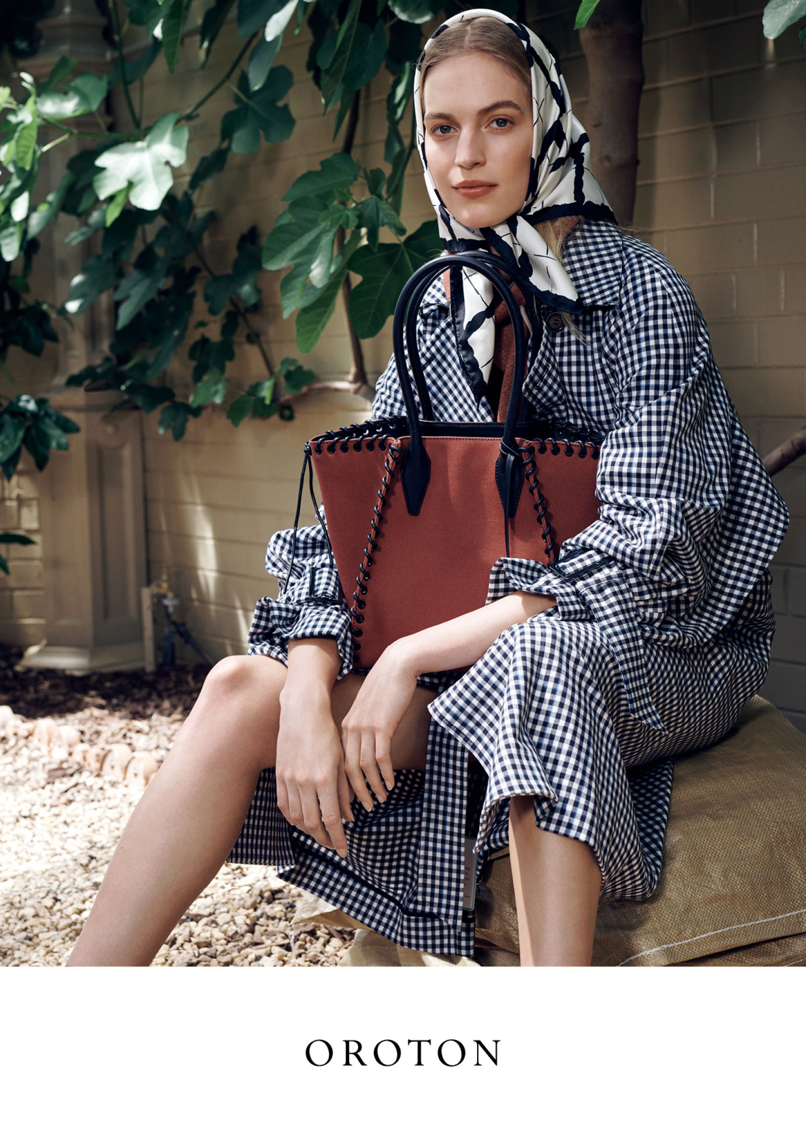 A fair-skinned woman sits outdoors with a reddish-brown leather tote bag, wearing a gingham dress and a patterned scarf on her head.
