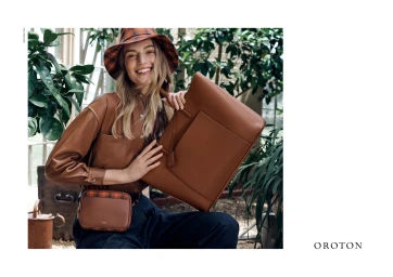 Smiling model holding a large brown leather bag wearing a matching plaid bucket hat and shirt in a lush greenhouse setting.