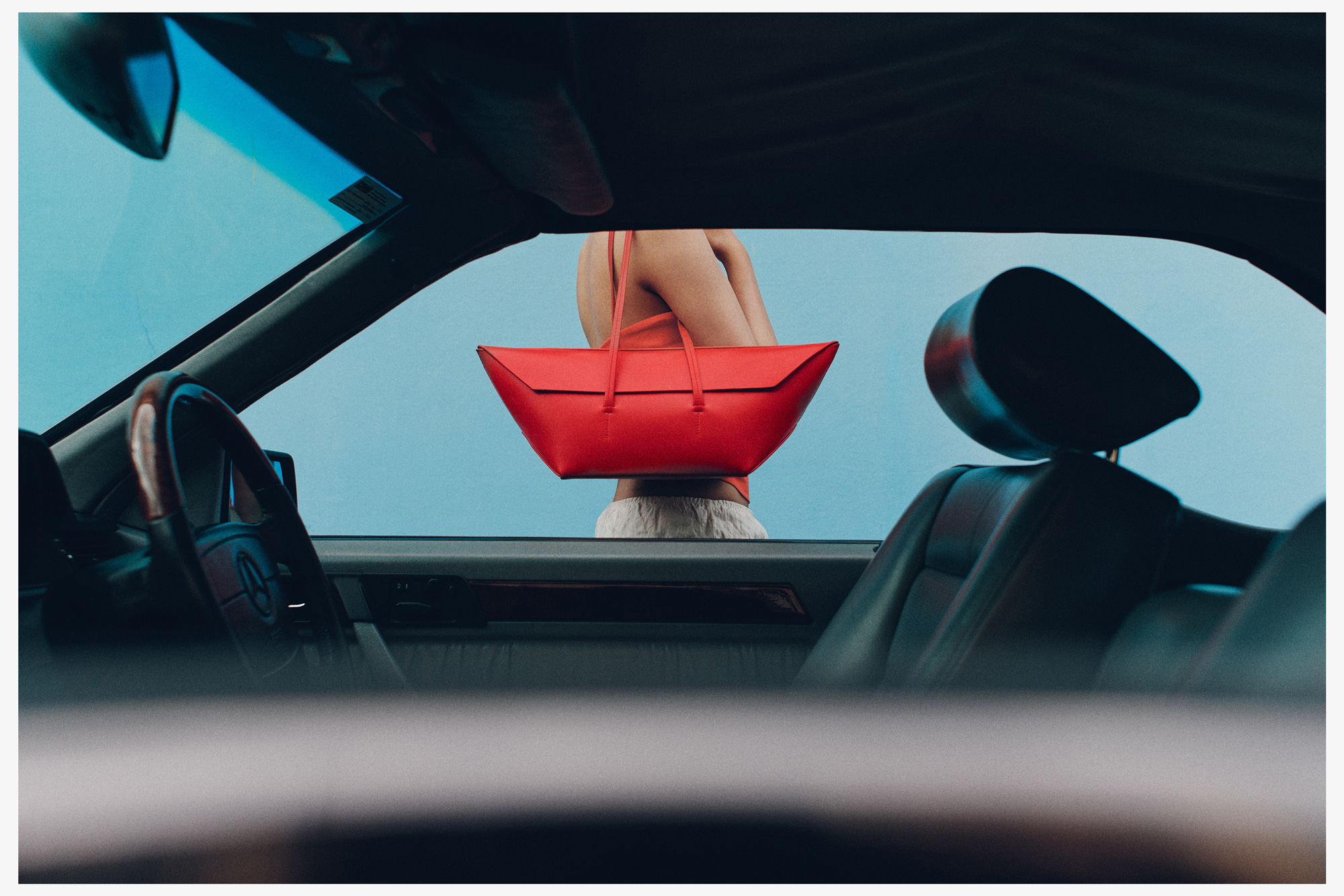 Red leather handbag held by a woman viewed through the open window of a car interior against a blue backdrop