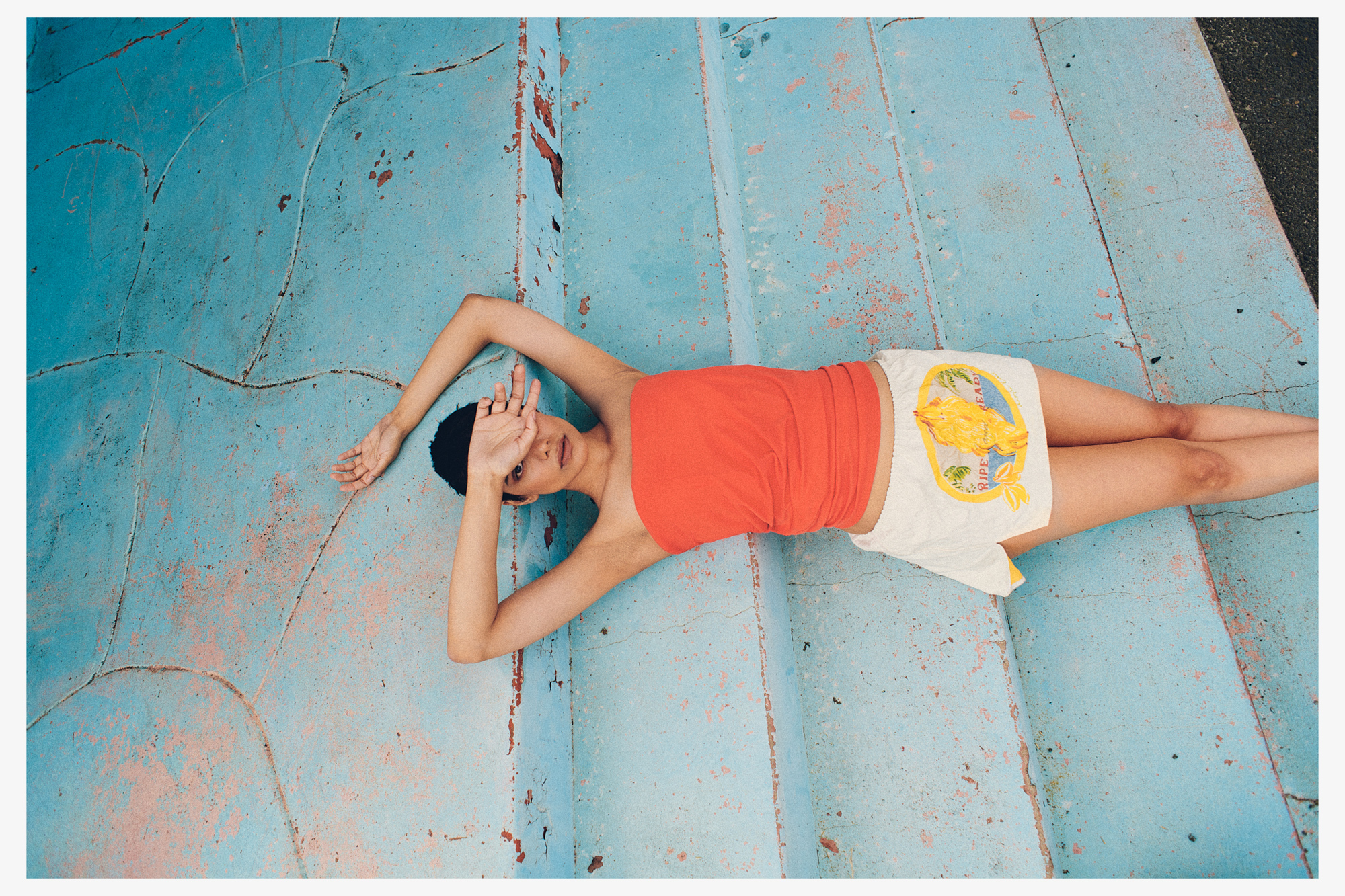 A woman reclines on worn, light blue steps, wearing a strapless orange top and patterned shorts, hands shielding her face from the sun.