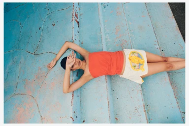 Model lying on weathered blue concrete steps wearing an orange top and patterned shorts for Christopher Esber.