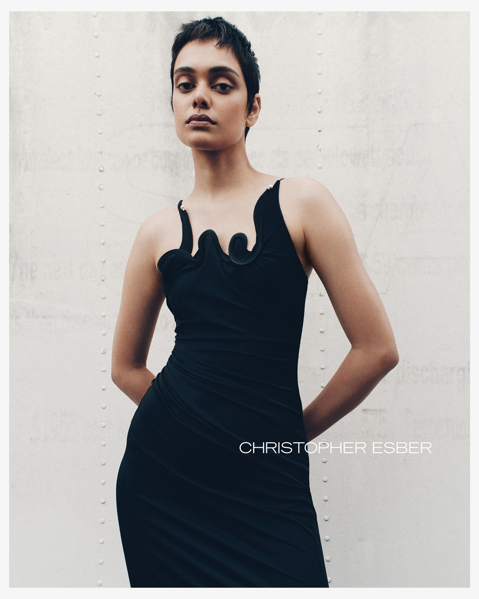 A fashionable woman with short dark hair poses in a black dress against a riveted white wall in a studio fashion shot.