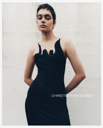 Model with short dark hair wearing a fitted black dress with sculptural neckline details against a metallic wall
