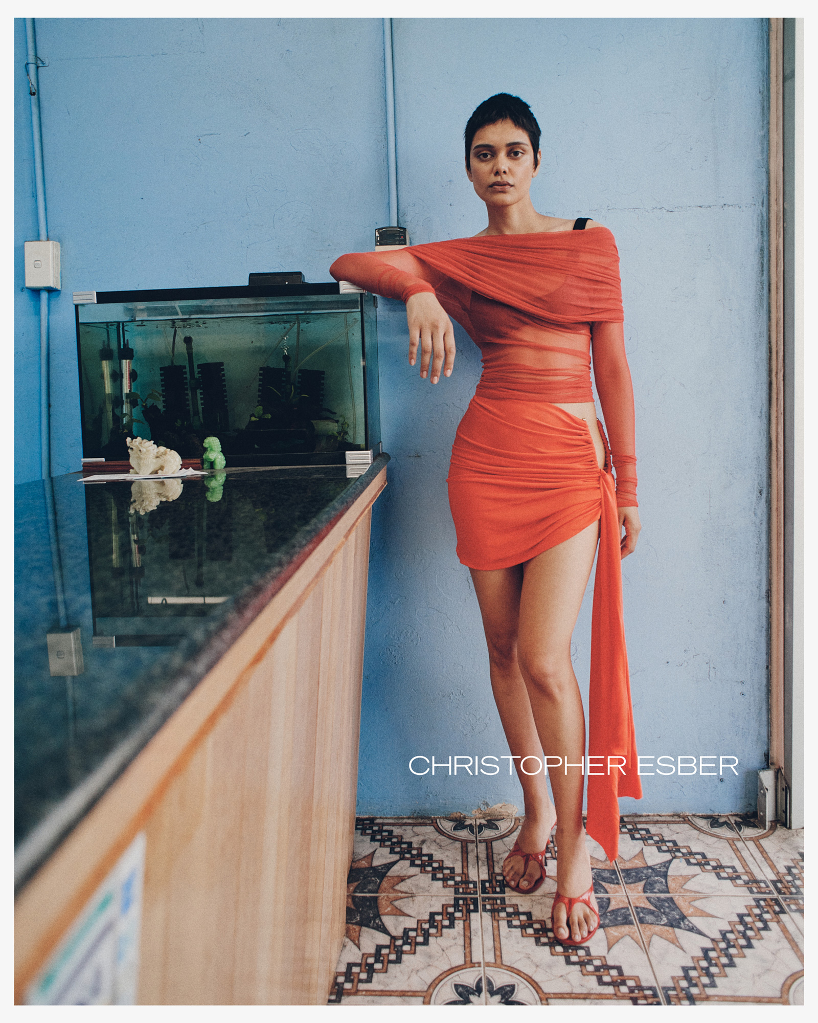 Fashion shot of an adult female wearing a red dress in a blue interior, posing near a fish tank and tile floor, with a designer logo.