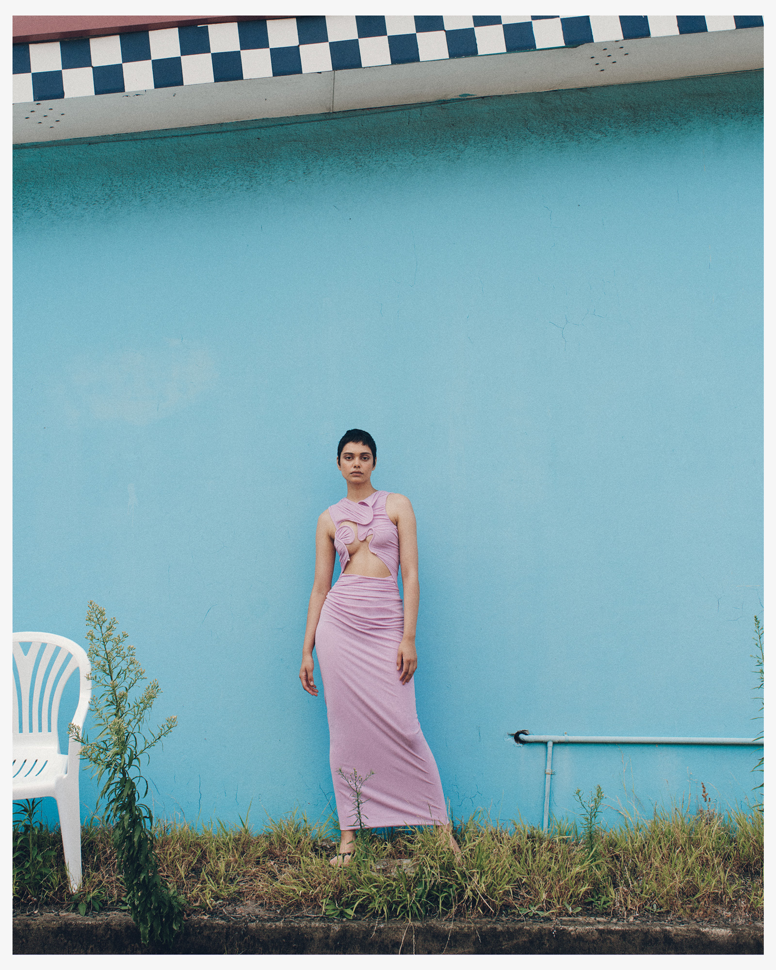 Woman in lavender dress poses against a blue wall with checkerboard trim, showcasing fashion and style in an outdoor setting with weeds.