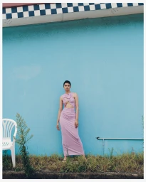 Model wearing a cut-out pink dress standing against a vibrant blue wall with a white plastic chair and overgrown weeds.