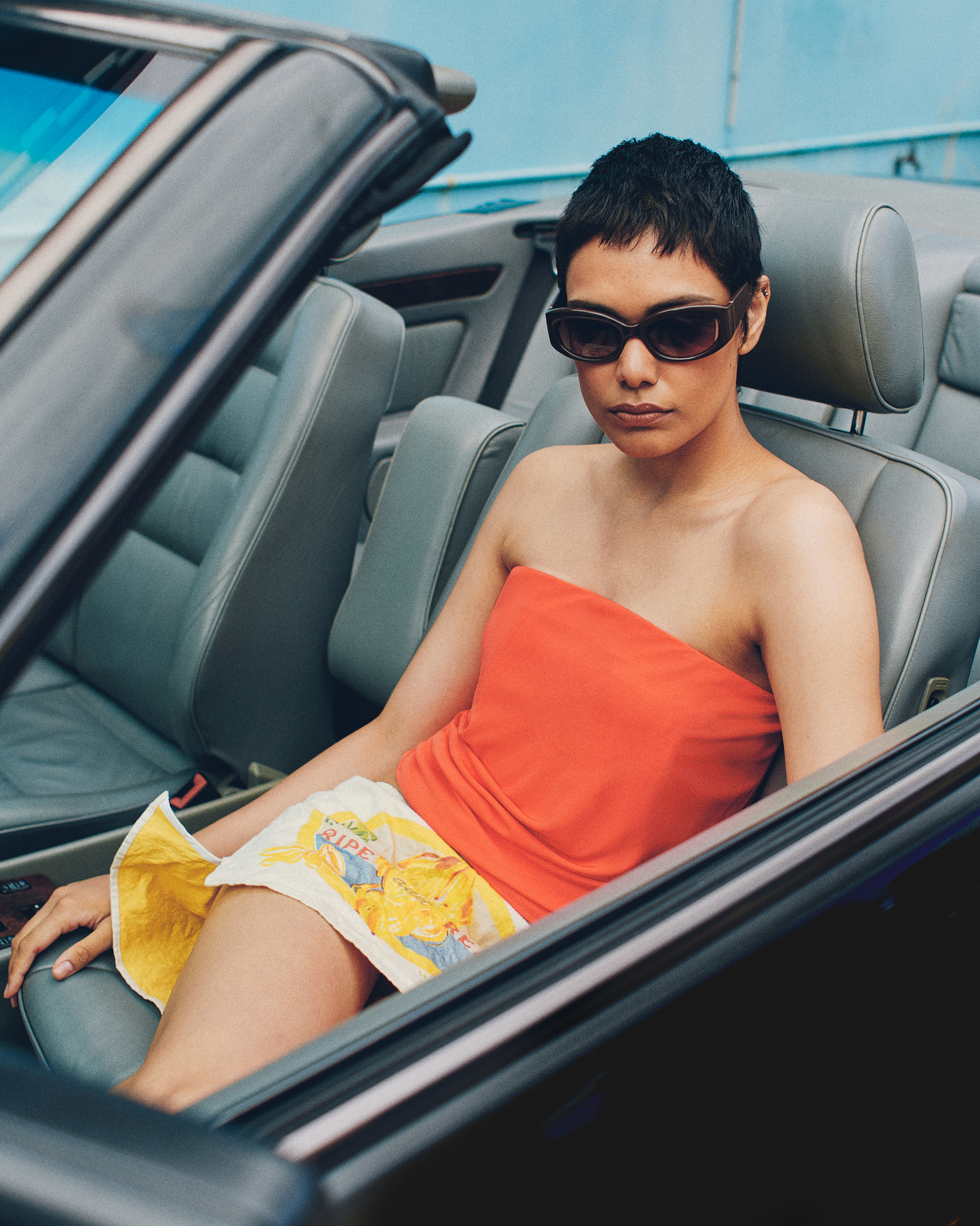 Model with short dark hair wearing sunglasses and an orange strapless top sitting inside a convertible car