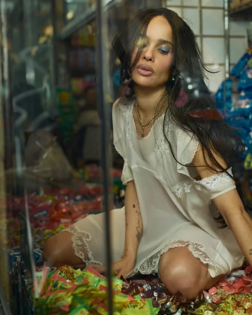 Adult female with blue eyeshadow kneels in a candy store, wearing a vintage nightgown, with bags of wrapped candy around her.