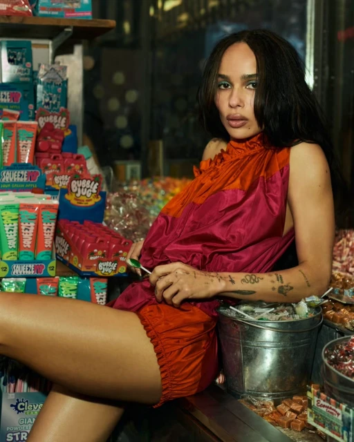Adult female wearing a pink and orange dress holding a lollipop in a candy store, with displays of sweets and treats.