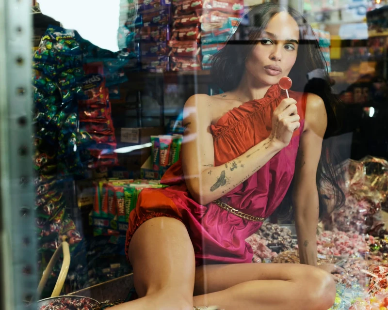 Zoë Kravitz poses in a red dress in a store, holding a lollipop with candy in the background, medium shot, eye-level angle.