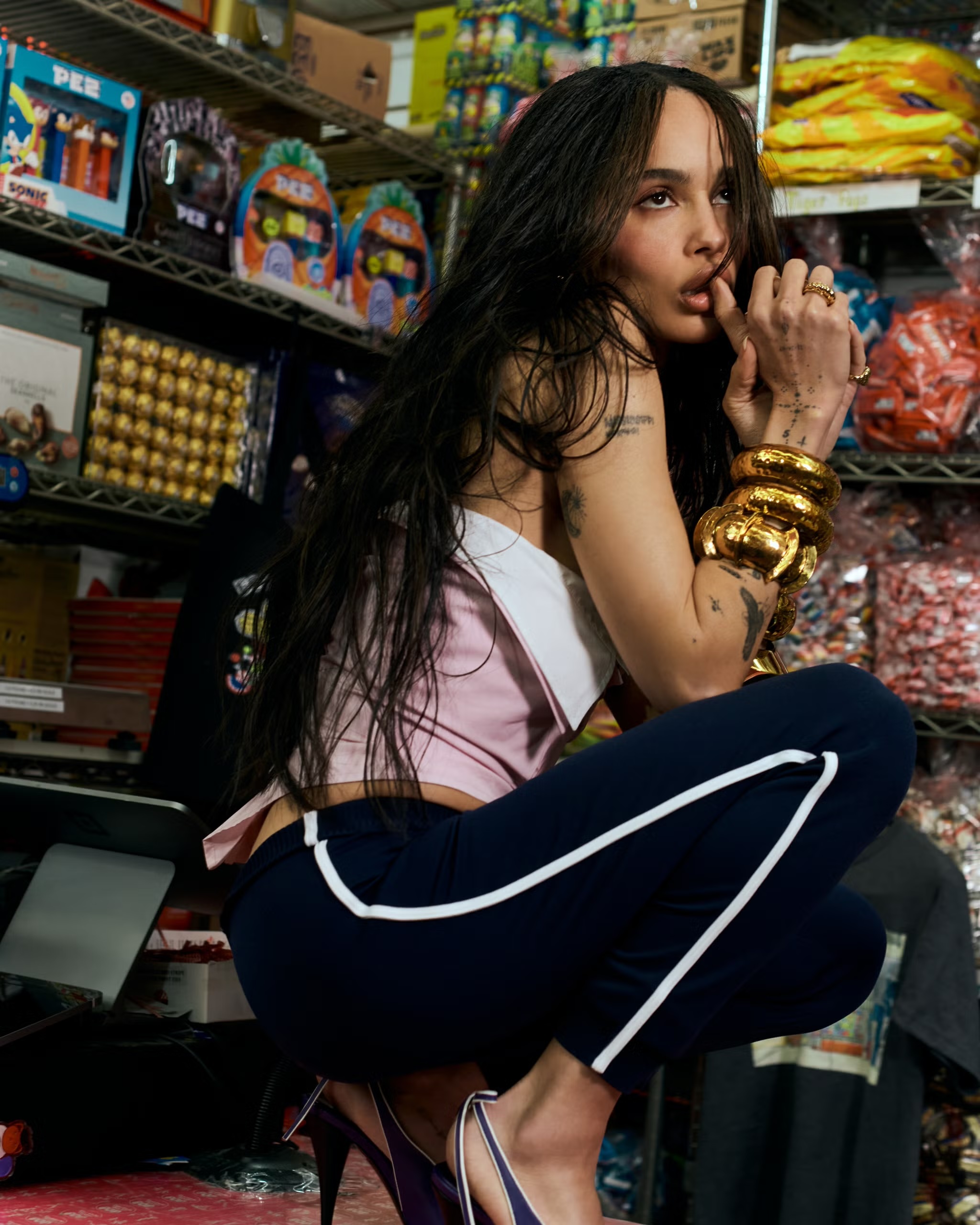 A woman with long hair and gold bracelets poses in a crouch with shelves filled with candy and collectibles in the background.