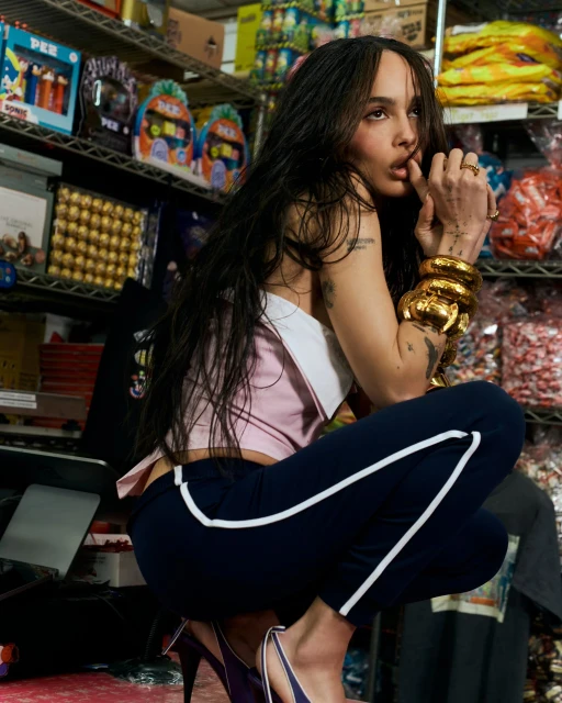 A woman with long hair and gold bracelets poses in a crouch with shelves filled with candy and collectibles in the background.