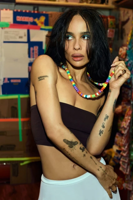 A woman with dark hair poses with a beaded necklace in front of shelves stocked with food products and packaging, indoor setting.