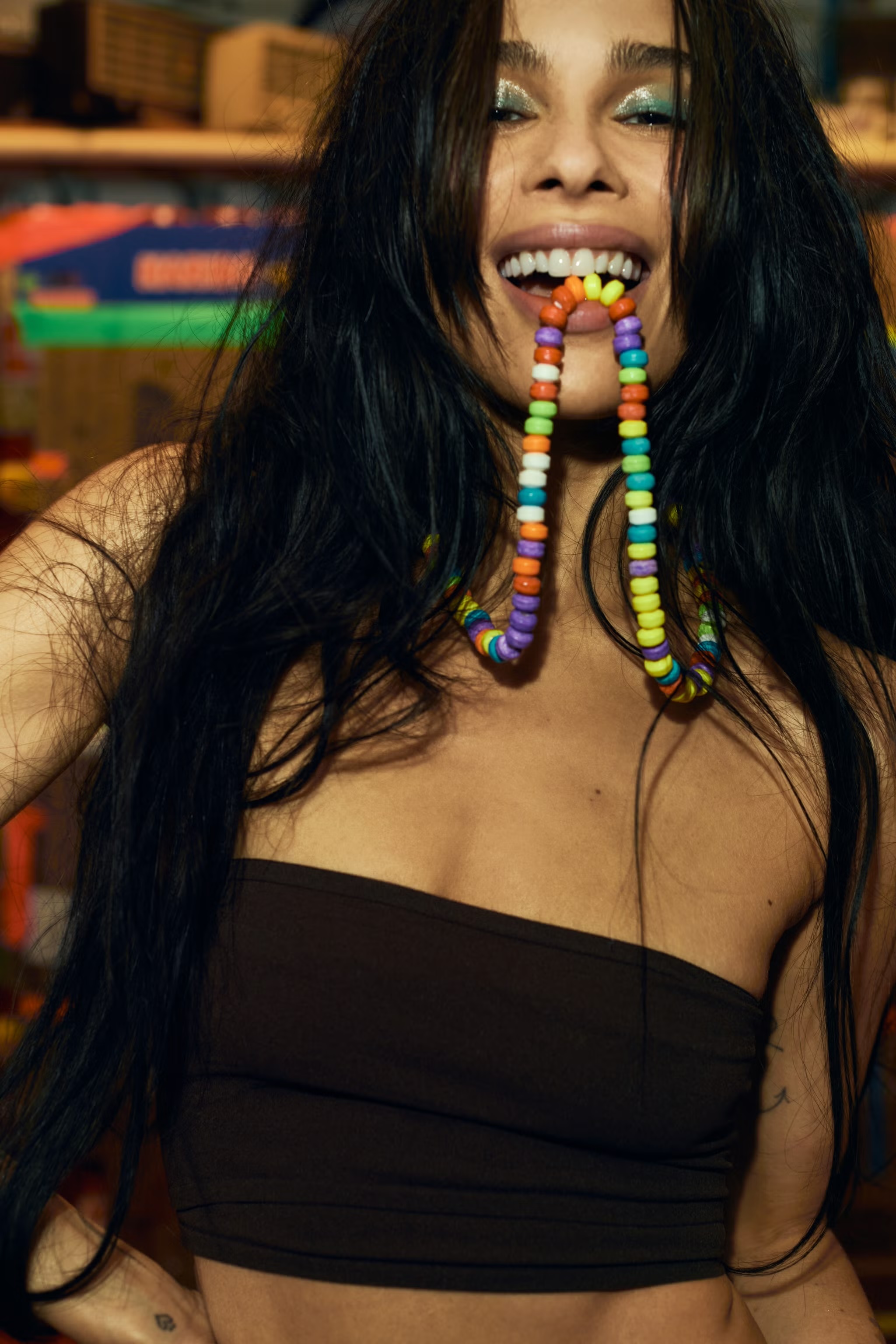 Portrait of a young adult woman with long dark hair, smiling with multicolored candy necklace in her mouth, wearing a brown tube top.