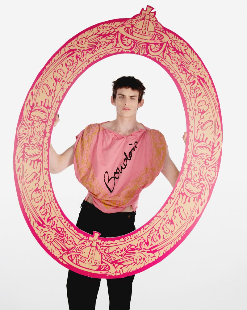 Male fashion model in a pink top and black pants poses with an ornate pink and beige frame on a white background in a fashion editorial.