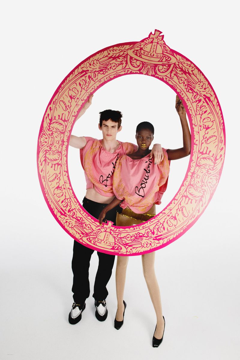 Two fashion models in pink logo shirts pose within a giant decorative frame on a white background in a studio setting, full shot.