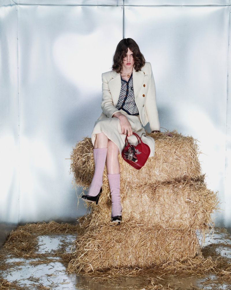 An adult female model sits on a hay bale in a studio. She wears a cream suit, pink leg warmers, and carries a red heart-shaped bag.