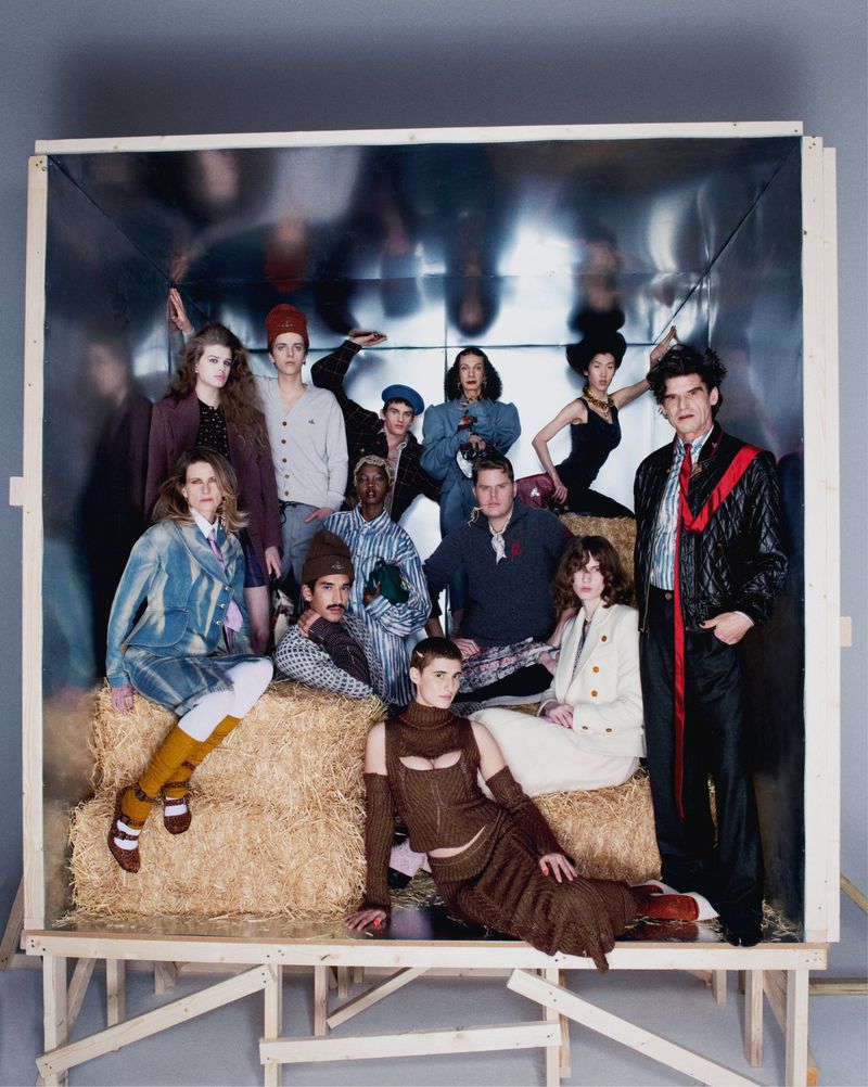 Group of fashionable people posing in a large wooden box with hay bales. Models showcase unique clothing styles in a studio setting.