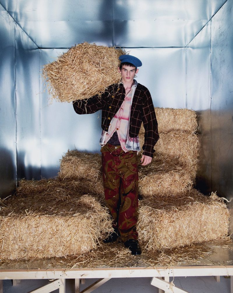 A young adult man in a beret and patterned outfit carries a bale of hay on his shoulder in a photo studio setting with silver walls.