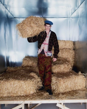 A young adult man in a beret and patterned outfit carries a bale of hay on his shoulder in a photo studio setting with silver walls.