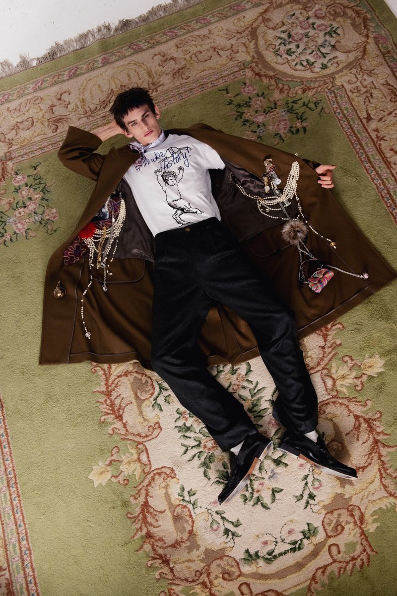 Young adult male in a white tee shirt and dark pants lying on a patterned carpet with a brown coat, jewelry, and accessories. 