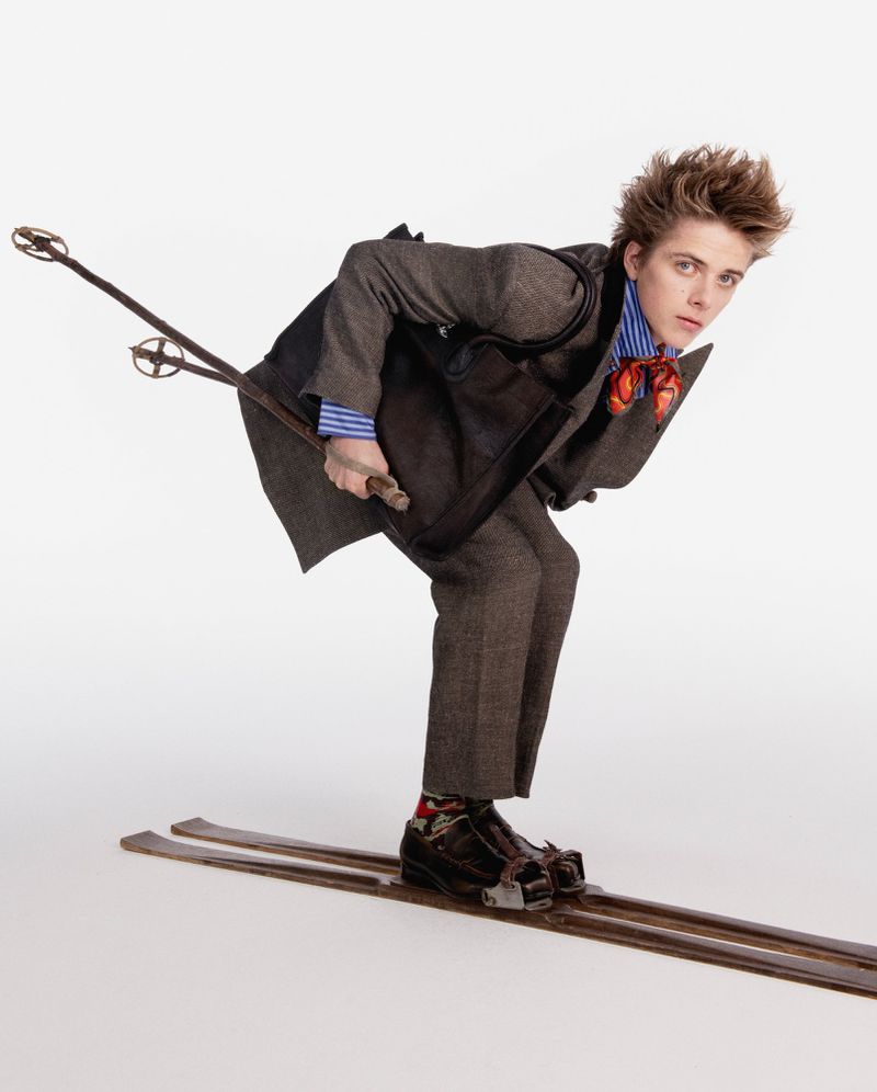 Male fashion model in tweed suit poses on skis with ski poles in a studio shot against white backdrop, showcasing vintage style.