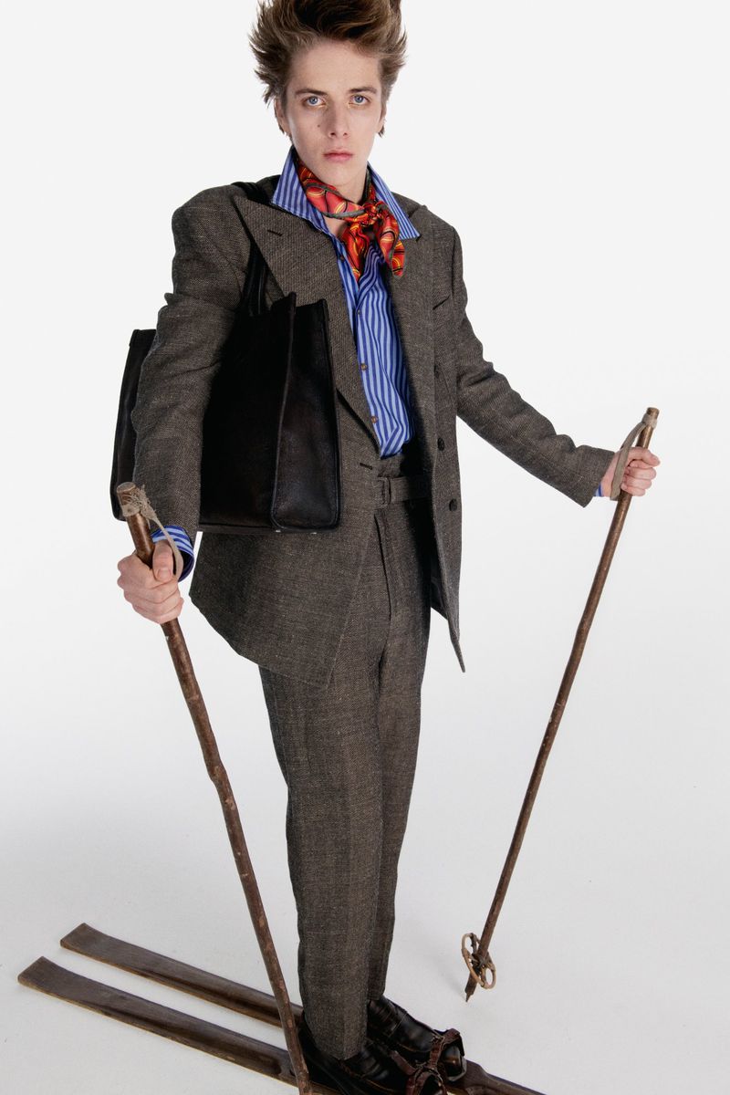 Person in a tweed suit, blue striped shirt, and red scarf standing on skis and holding poles against a white backdrop. Fashion shoot.