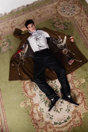 Model lying on an ornate patterned green rug wearing a long brown coat open over a printed graphic t-shirt and black trousers.