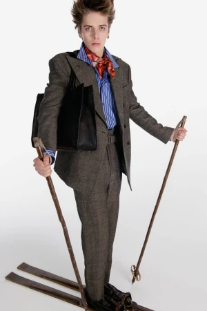 Model posing in a tailored grey tweed suit with vintage wooden skis, poles, and a black tote bag on a white studio background.