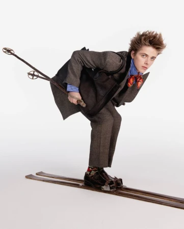 Model in a brown tailored suit crouching on vintage wooden skis with poles against a bright white background.