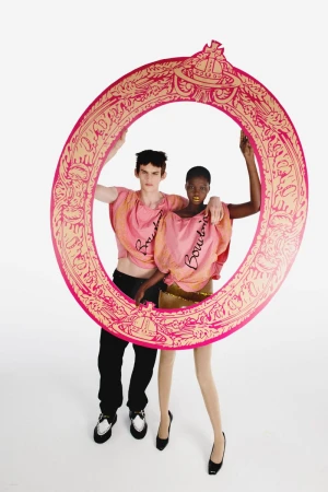 Two models standing behind a large pink and gold ornate oval cutout frame in matching pink draped tops against a white studio background.