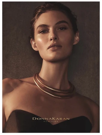 Close-up studio shot of a woman wearing a gold necklace and black strapless top, bathed in soft lighting against a dark grey backdrop.
