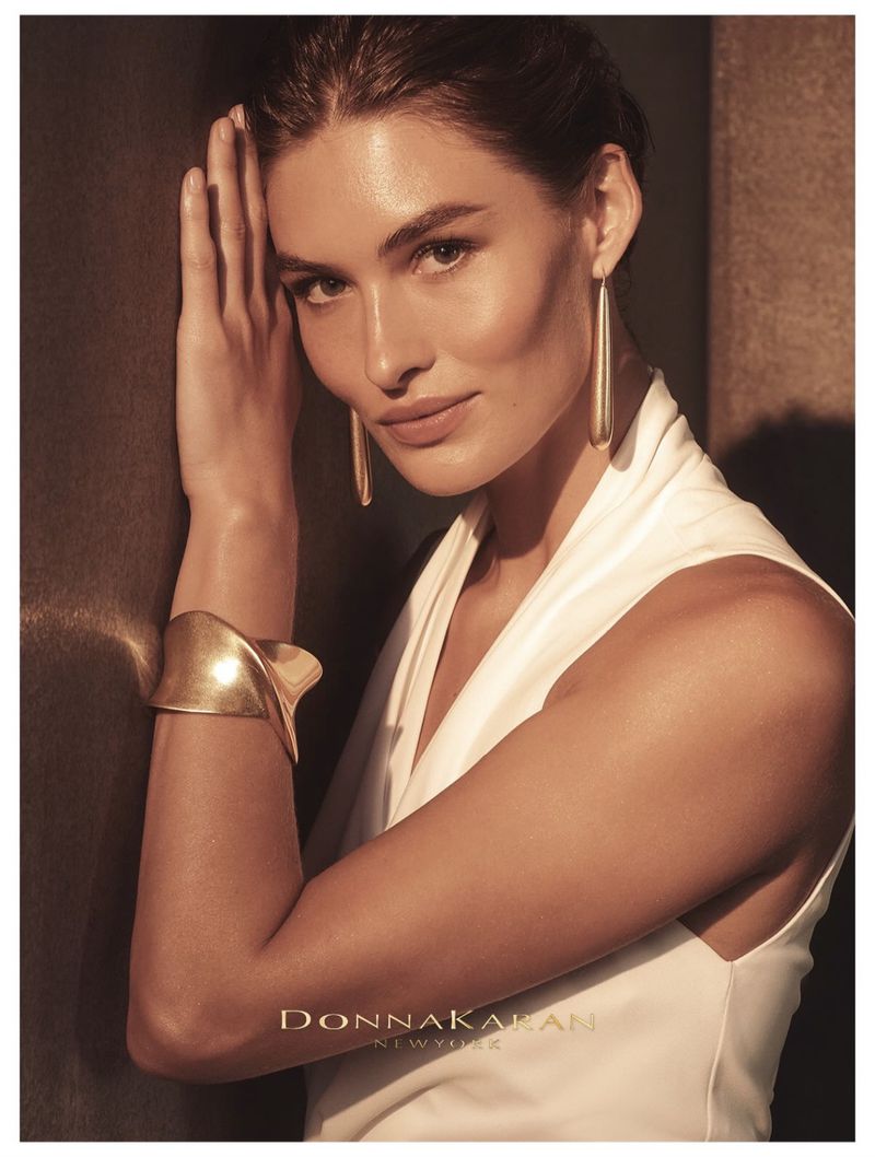 Elegant portrait of an adult woman wearing long gold earrings and a gold cuff bracelet, leaning against a dark wall in soft light.
