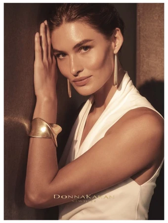 Elegant portrait of an adult woman wearing long gold earrings and a gold cuff bracelet, leaning against a dark wall in soft light.