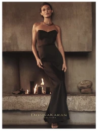 Model Grace Elizabeth wearing a black strapless evening gown and gold choker necklace standing beside a fireplace for Donna Karan Holiday 2025.