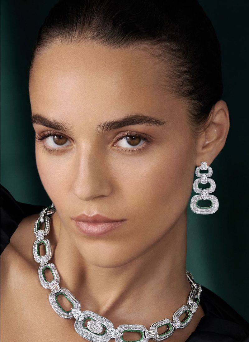 Close-up of a woman with light skin wearing a diamond and emerald necklace and earrings against a dark green background.