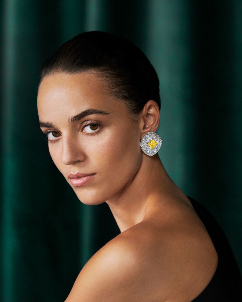 Close-up studio shot of a woman with dark hair and tan skin, wearing a black dress and diamond earrings with yellow gemstone.
