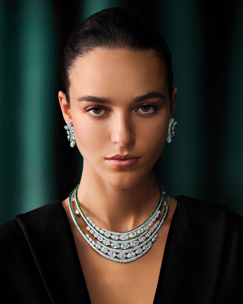 Close-up of a woman modeling diamond jewelry, including a multi-layered necklace and earrings, against a dark green background.