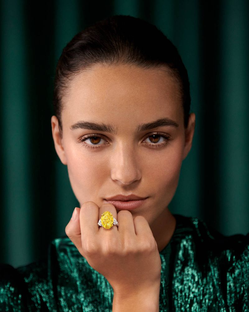 Woman poses indoors with a yellow diamond ring on her finger, wearing a green textured top, in a close-up studio shot against a green backdrop.