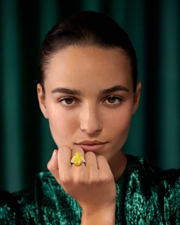 Woman poses indoors with a yellow diamond ring on her finger, wearing a green textured top, in a close-up studio shot against a green backdrop.