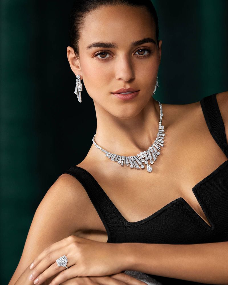 Elegant woman with dark hair poses wearing a black sleeveless dress, diamond necklace, ring, and earrings against a dark green background.