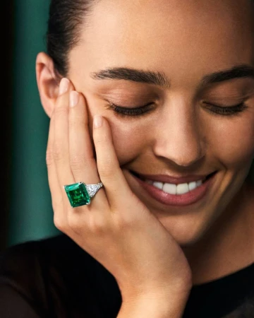 Close-up of a woman's face and hand featuring a large emerald ring, set against a dark teal background.