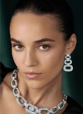 Model Emm Arruda wearing a diamond and emerald chain-link necklace and tiered drop earring against a dark green backdrop.
