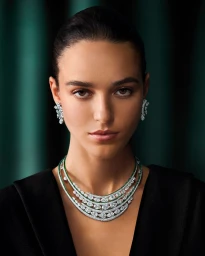 Model Emm Arruda wearing a multi-tiered Graff diamond and emerald necklace and earrings against a dark green backdrop.