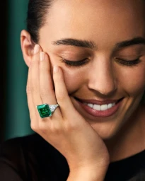 Close-up of a smiling model resting her hand on her cheek, showcasing a large emerald-cut green gemstone ring.