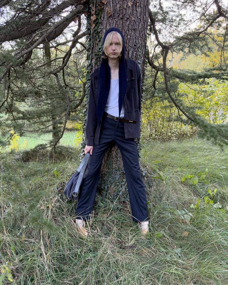 A young adult with blond hair in a dark coat and jeans poses against a tree in a grassy, wooded area under natural daylight, holding a bag.