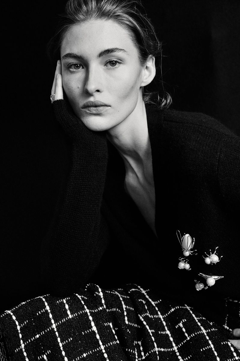 Black-and-white portrait of a fair-skinned woman with neutral expression, wearing a dark cardigan with bug brooches and a patterned skirt.