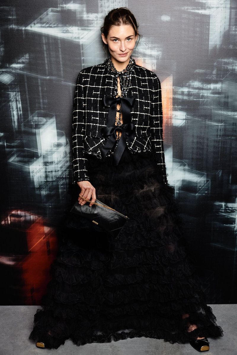 Fashionable woman poses in a black ruffled skirt and patterned jacket against a muted city backdrop, holding a stylish clutch.