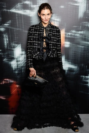 Fashionable woman poses in a black ruffled skirt and patterned jacket against a muted city backdrop, holding a stylish clutch.